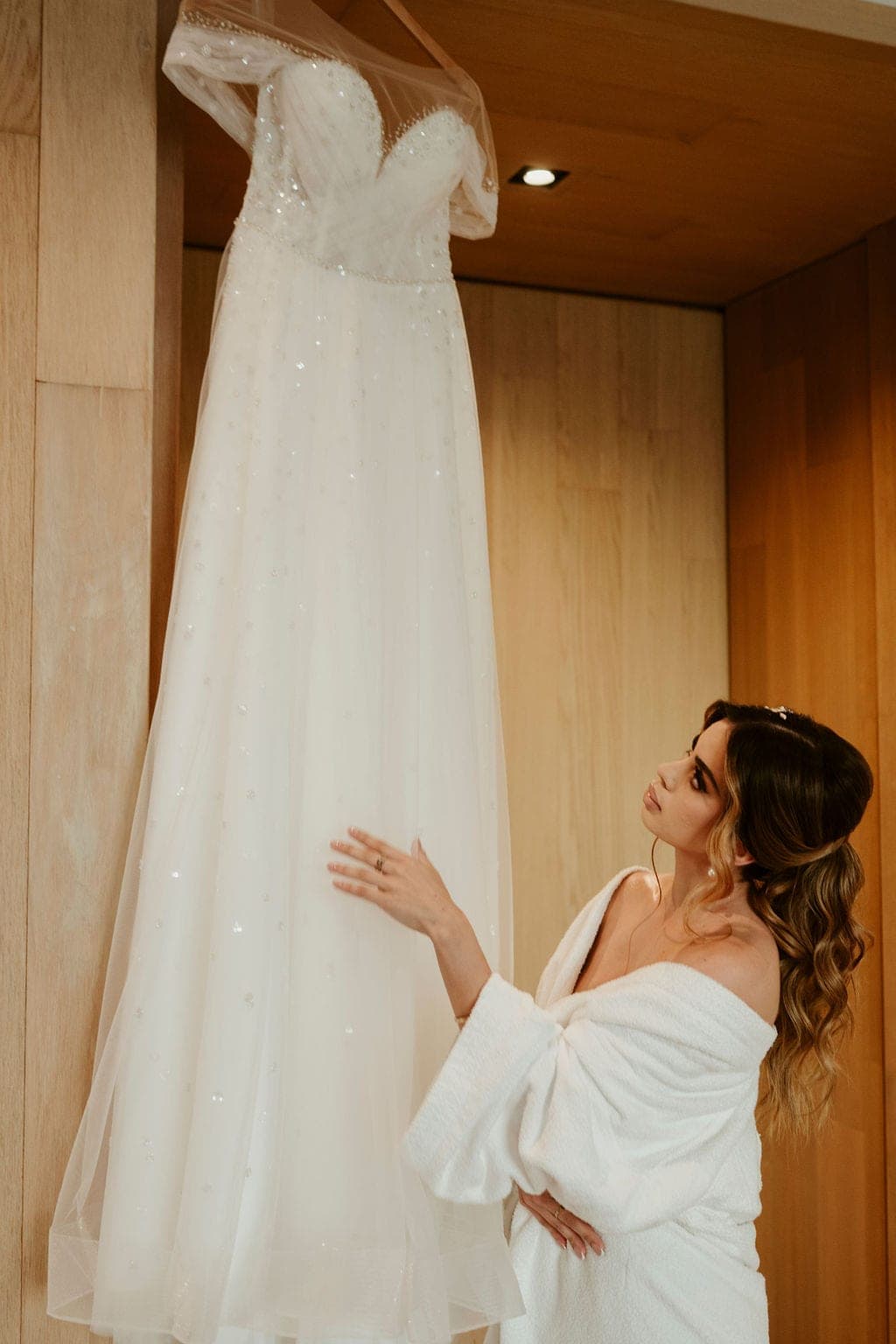 Novia en bata blanca tocando su vestido colgado en una habitación de madera