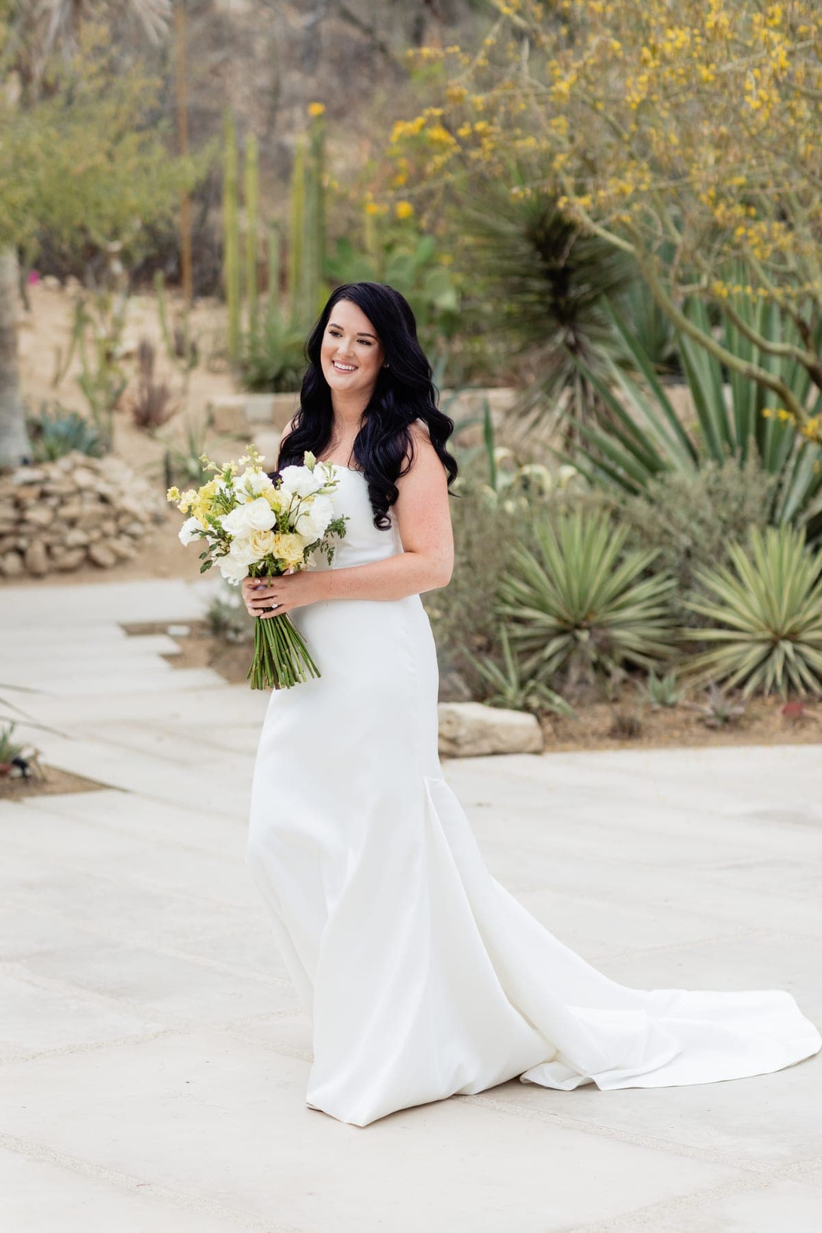 Novia de ondas largas y oscuras con un ramo blanco y amarillo en un jardín de cactáceas