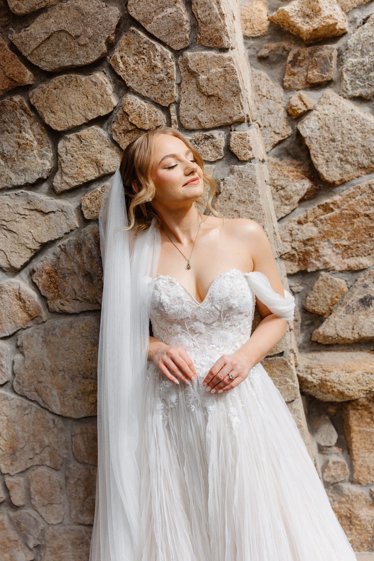 Novia con velo largo recargada en un muro de piedra, con los ojos cerrados al sol