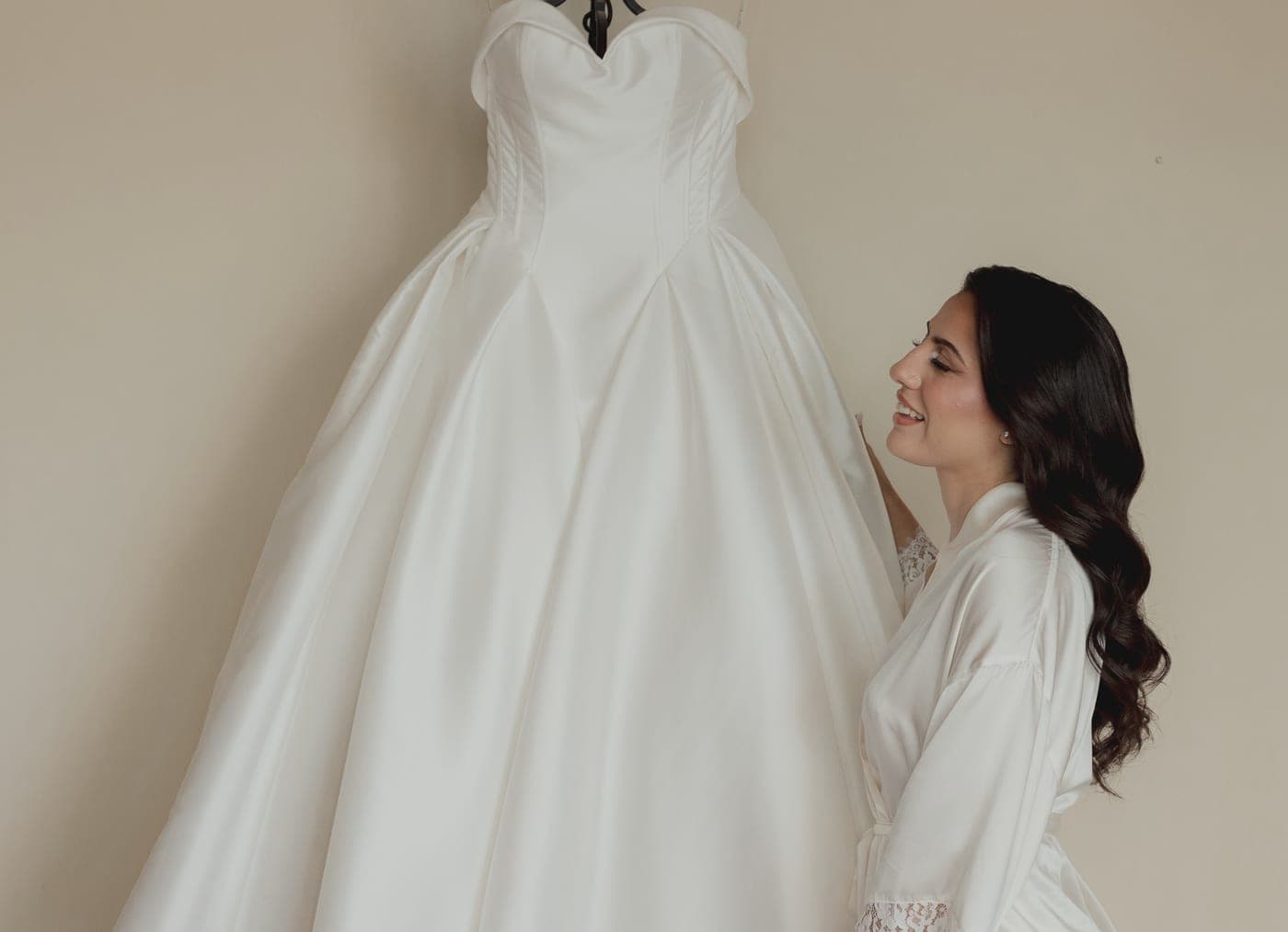 Novia en bata de satín sonriendo a su vestido colgado en la pared